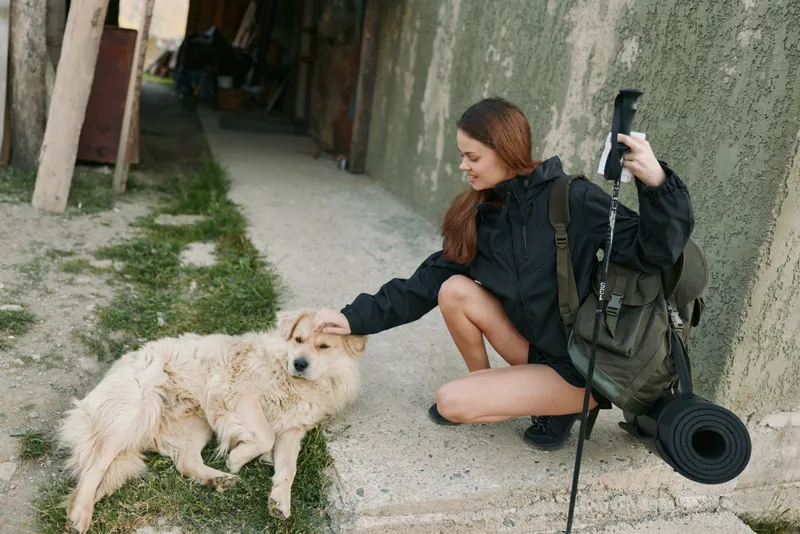 A young woman crouches beside a fluffy dog in a sunlit alley. She wears a dark jacket and backpack, with a rolled mat, sharing a calm, outdoor moment.