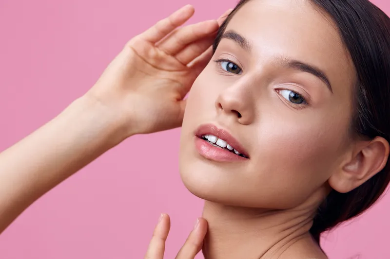 Young model with natural beauty showcasing clear skin, delicate features, and a soft expression against a pink background, representing skincare and self care