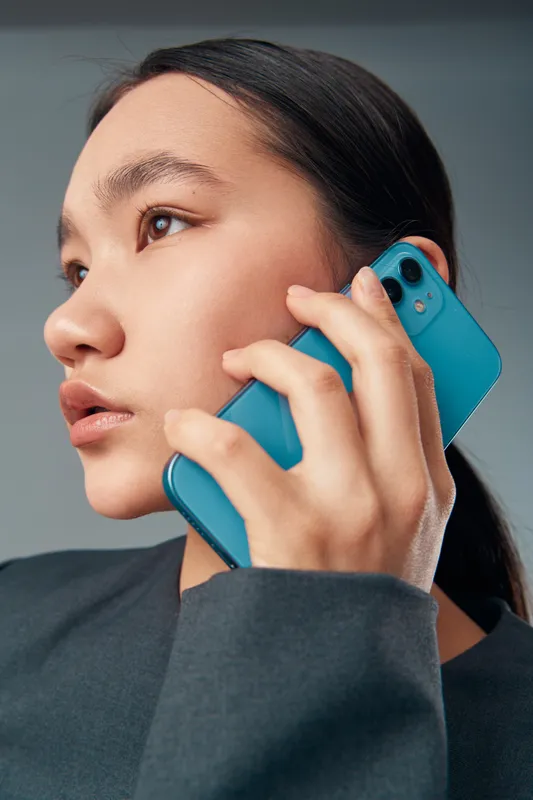 Young woman holding blue smartphone close to ear for call or conversation with serious expression. Closeup portrait with natural light and minimalistic style for communication or technology.