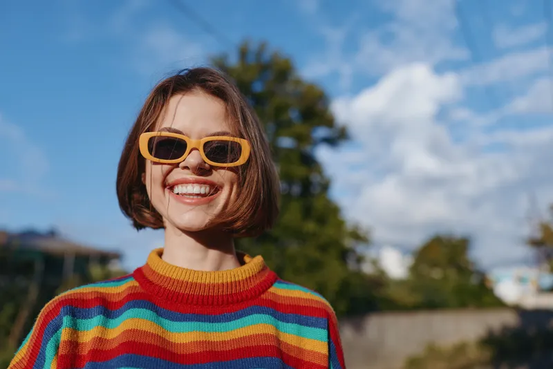 Woman smiling in rainbow sweater and yellow sunglasses outdoors, short hair portrait under blue sky, joyful young adult, casual outfit, sunny lifestyle travel vibe and playful fashion mood.