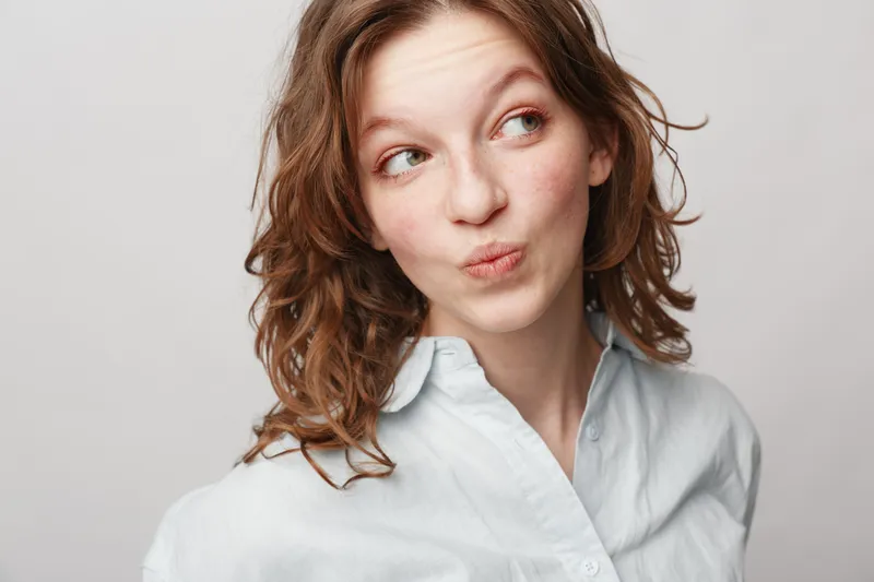 Young woman with curly hair wearing white shirt looks sideways with playful expression on light background. Concept of casual style, youth, and natural beauty portrait.