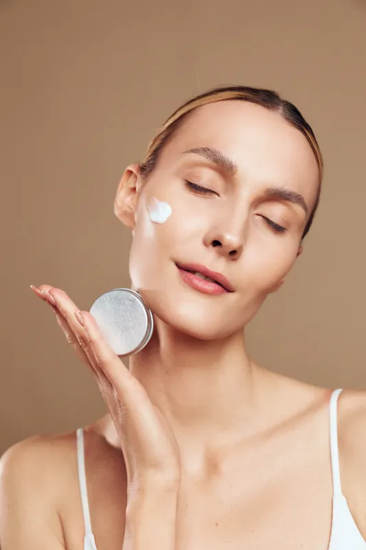 A young woman with clear skin applies white cream to her cheek while holding a round cosmetic container near her face. The neutral background emphasizes skincare and beauty care concept.