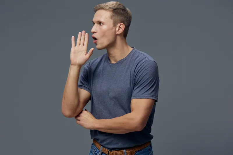 Confident man in casual blue t-shirt and jeans gestures as if telling a secret or warning, standing against a solid gray background. He appears engaged and expressive, suitable for concepts of communi