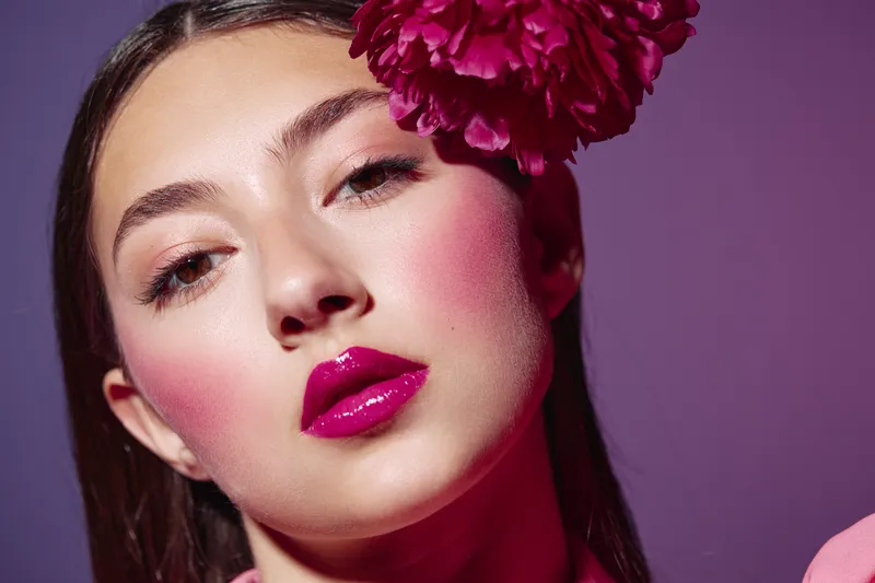 Young woman with glossy bright pink lips and bold blush wearing a large pink flower on head. Close-up on purple background. Concept of beauty fashion creative style and makeup.