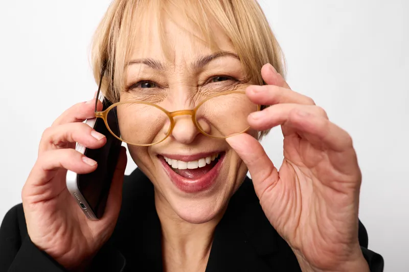 Senior woman, laughing, holding glasses, talking on phone, closeup portrait, indoor bright light, cheerful expression, relaxed hands, white background