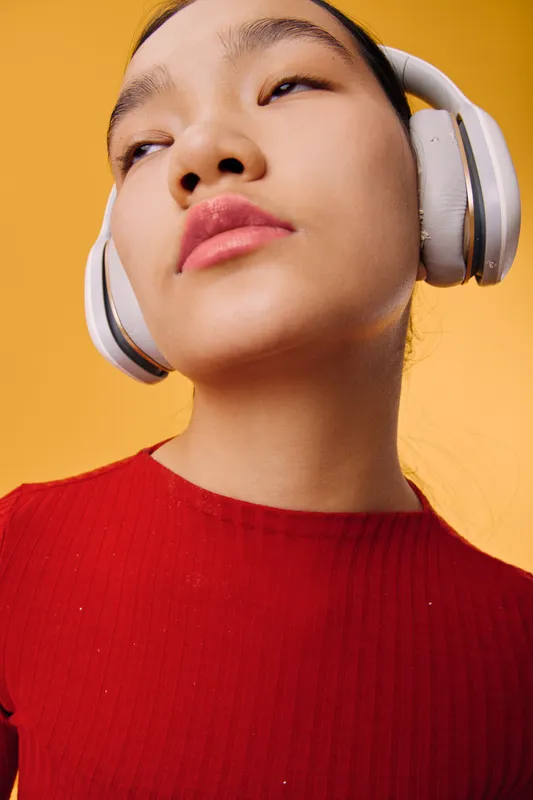 Young woman wearing white headphones and red top listening to music with eyes closed on yellow background. Closeup portrait with serene expression and modern lifestyle vibe.