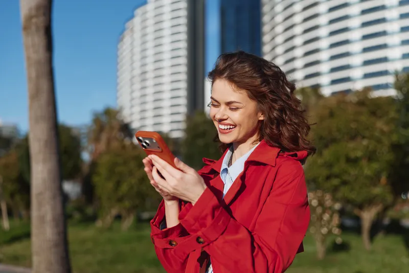 Young woman in a red coat smiles while using a smartphone in a sunny city park, capturing a candid outdoor lifestyle moment with brown hair and casual clothing, open space around