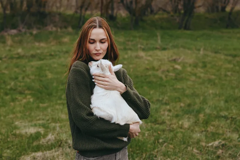 Young woman in a green sweater gently holds a white baby goat outdoors in a grassy field during a calm lifestyle moment.