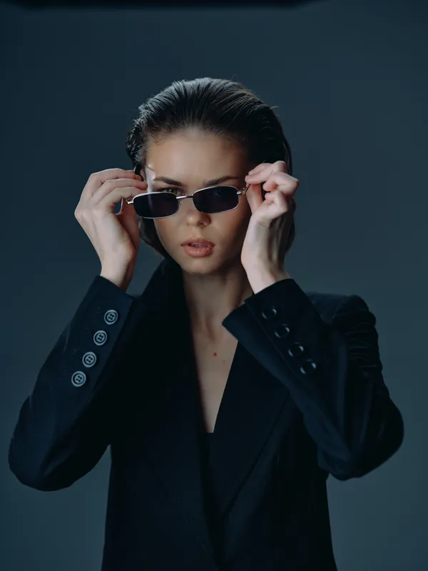 Woman wearing sunglasses and black blazer with serious expression in moody lighting