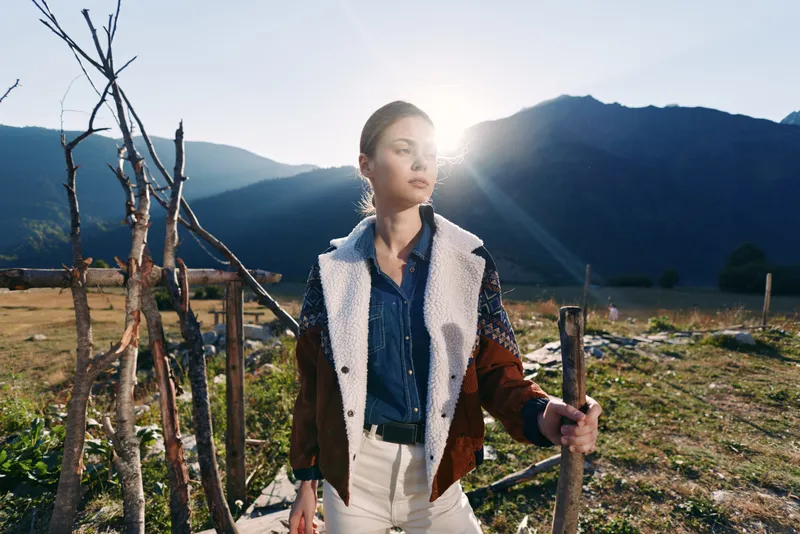 Woman in denim shirt and shearling jacket stands by a wooden fence in rural mountains landscape at sunrise with sunlight backlight and confident gaze, casual outdoor portrait.