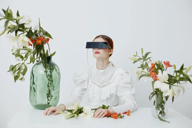 A woman in a white vintage dress with futuristic glasses surrounded by flowers in a minimalist setting symbolizes harmony between nature and technology