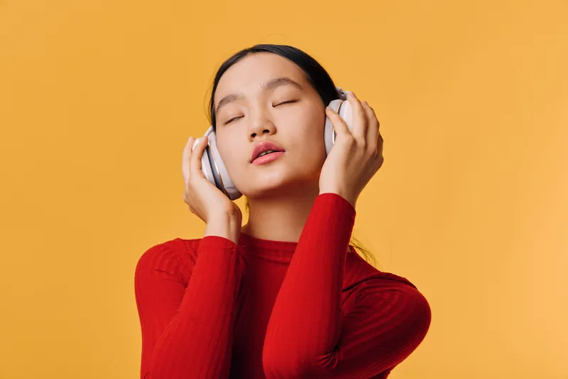 Woman with closed eyes wearing headphones and red sweater listening to music. Relaxation, leisure, and enjoyment with yellow background and calm expression on face in studio.