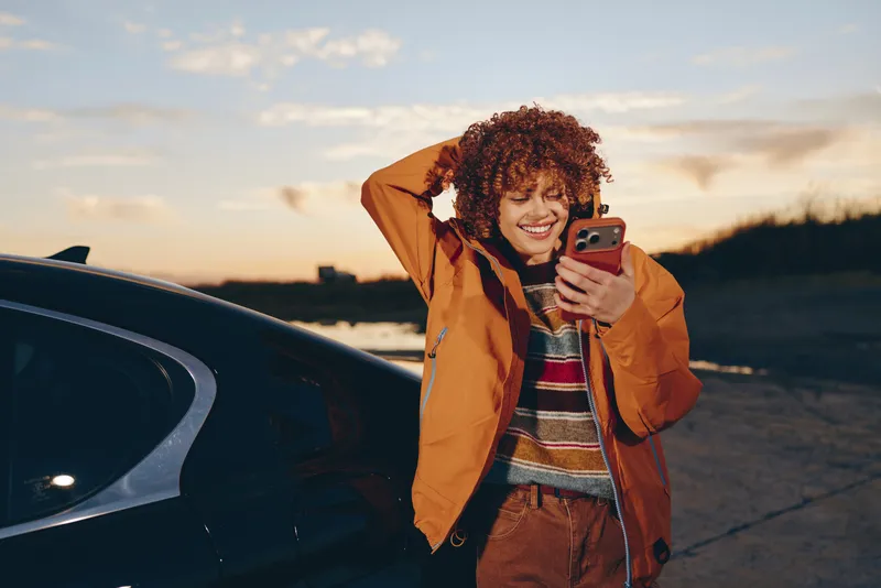Smiling woman with curly hair in orange jacket stands by a car at sunset holding a smartphone and chatting outdoors lifestyle photo with warm golden light roadside moment roadtrip