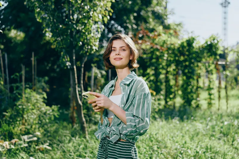 Young woman in a light jacket stands in a sunny garden, holding a small green fruit and offering a calm, cheerful smile.