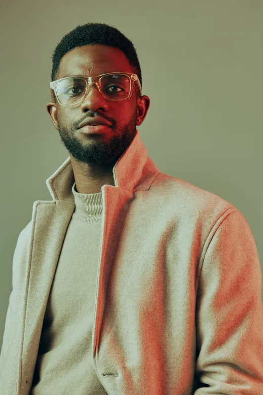 Stylish young Black man wearing glasses and a light beige coat, posing against a modern, softly colored background, conveying confidence and sophistication