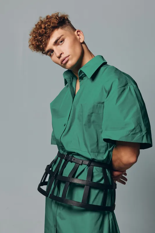 Young man with curly hair wearing an oversized green shirt and black waist cage belt posing against gray background. Fashion, style and modern outfit concept with confident expression.