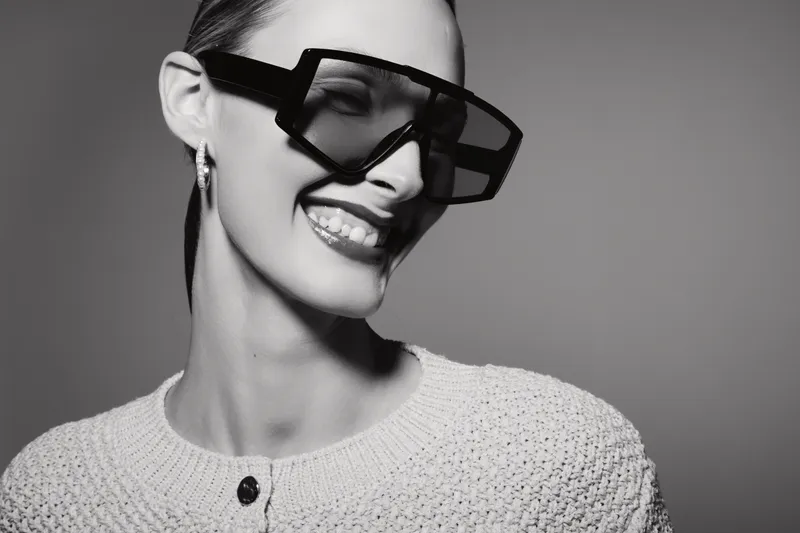 Smiling young woman with pale skin and dark hair wearing oversized geometric sunglasses and knitted sweater. Studio portrait in black and white with modern fashion concept.