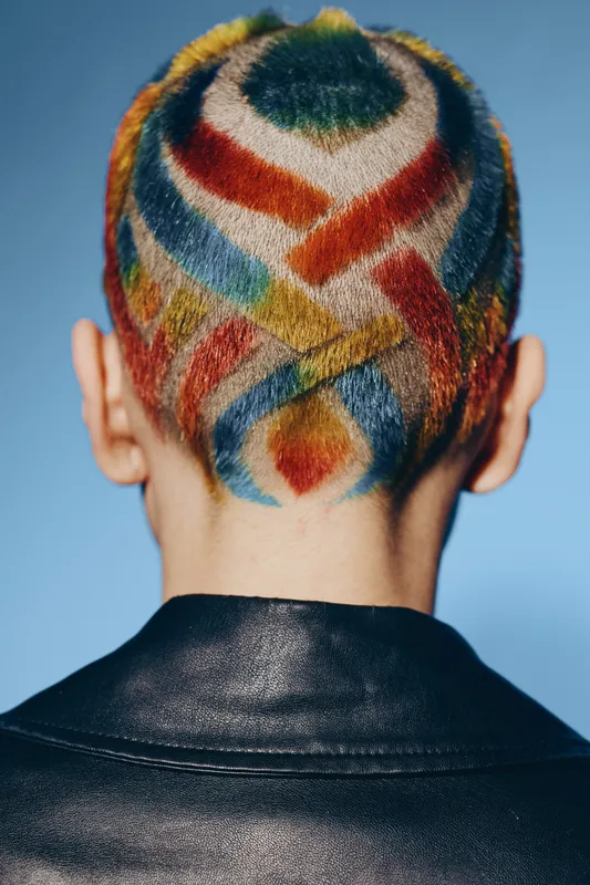 Woman with colorful geometric pattern shaved hairstyle viewed from behind, wearing a black leather jacket on blue background, bold and artistic look.