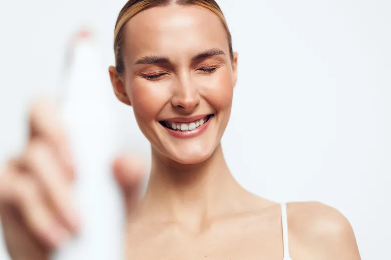 Young woman with closed eyes holds spray bottle and smiles gently while applying mist on face isolated on white background, skincare routine concept, fresh and natural beauty.