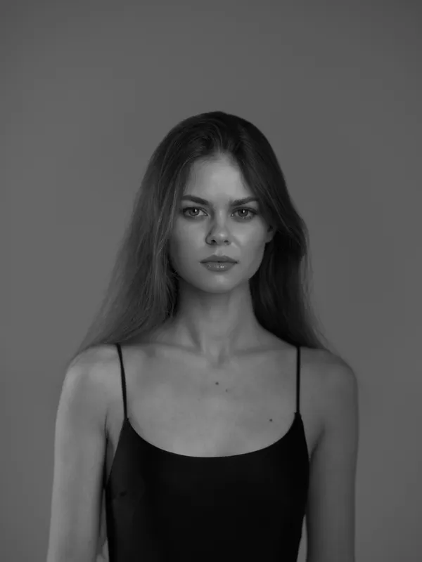 Black and white portrait of a serious woman with long hair wearing a black top with thin straps, isolated on gray background.