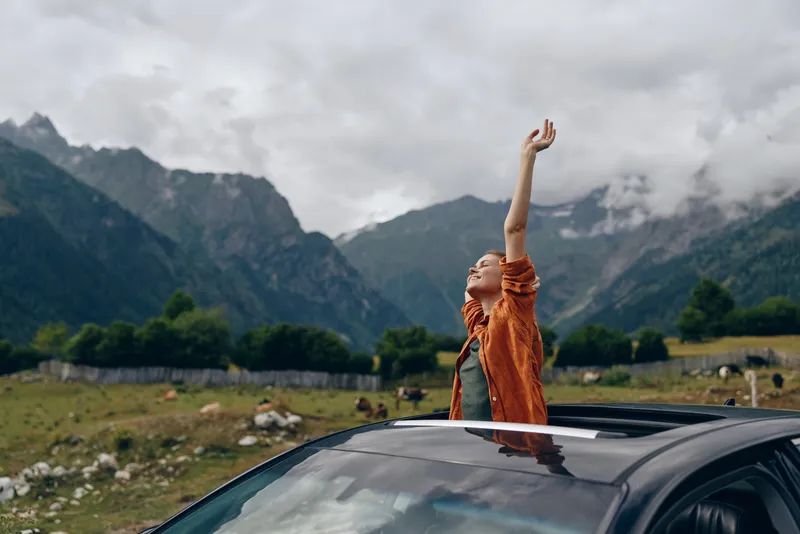 A person in an orange jacket stands through a car sunroof, raising an arm in the alpine landscape, expressing freedom and adventure amid mountains and open skies