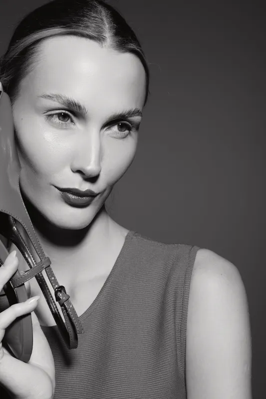 Close-up portrait of a smiling Caucasian woman with fair skin and slicked-back hair holding a handbag strap. The studio lighting highlights her natural beauty and confident expression.