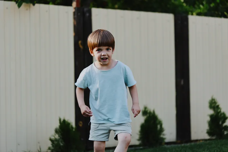 Active boy running joyfully in a garden, wearing a casual light blue t-shirt and shorts, surrounded by greenery and a wooden fence.