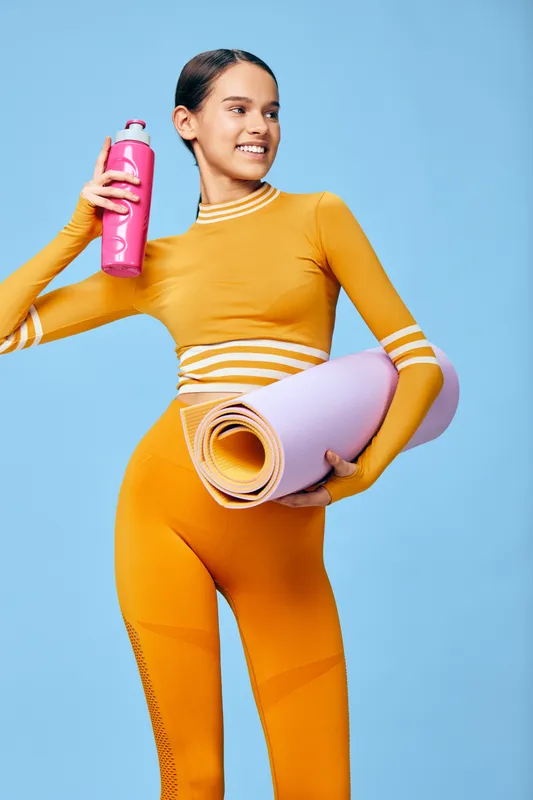 Young woman in orange activewear holding a rolled yoga mat and pink water bottle, smiling confidently against a bright blue studio background, fitness and sport lifestyle concept.