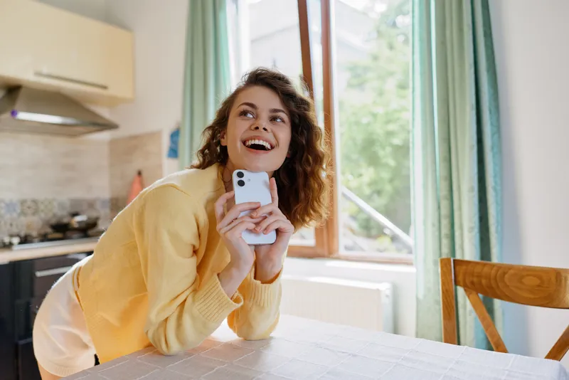 Bright woman at home lifestyle scene, smiling while holding a smartphone near a window, warm natural light, cozy kitchen, casual sweater, joyful everyday moment.