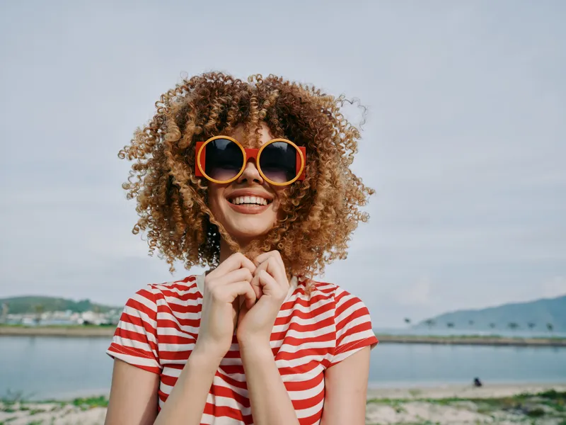 Bright portrait of a joyful woman with curly hair and oversized orange sunglasses, wearing a red striped tee by a sunny beach, radiating playful confidence.