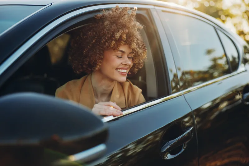 Smiling woman with curly hair in a car, window down, daylight highlighting her smile. Retro film color mood, casual lifestyle, travel vibe, warm, natural lighting.