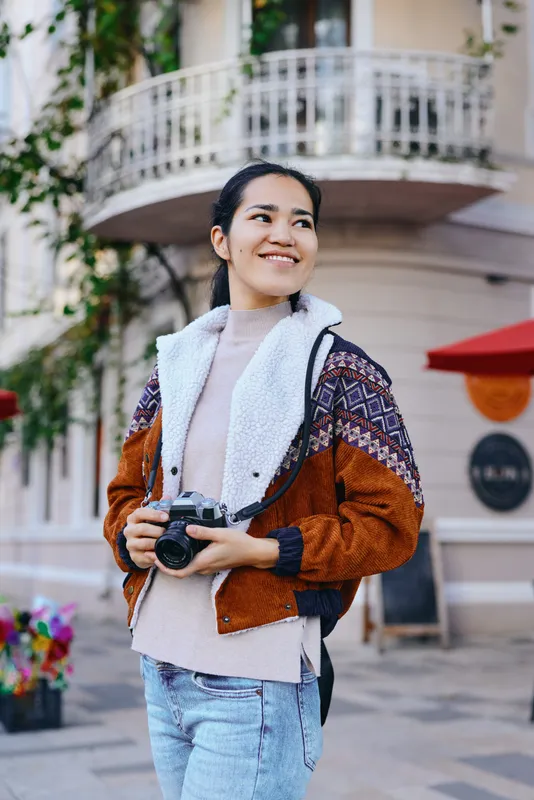 Camera, photographer exploring street life with a warm scarf and jacket, smiling woman walks outdoors as archives of travel style unfold in a sunny urban setting, capturing candid moments and
