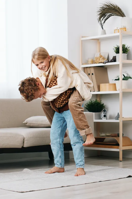 Two children playfully wrestle indoors, one perched on the other's back, smiling and laughing as they enjoy a cozy moment on the living room rug, radiating warmth.