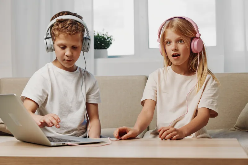 Two children wearing headphones use a laptop while sitting at a table in a bright living room, focused on digital learning and entertainment together.