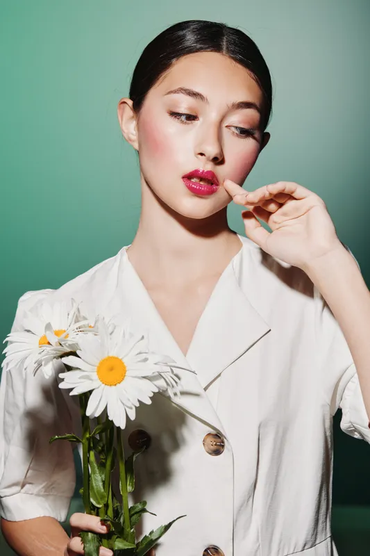 Young woman in white dress holds chamomile flowers while touching her face, posing creatively in front of green background. Concept of fashion, beauty, nature, and minimalism.