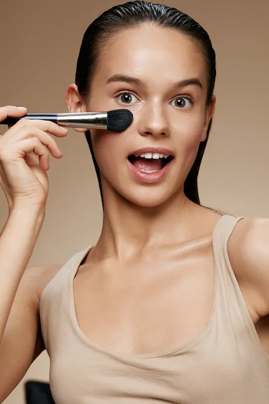 Cheerful young woman applying makeup with a brush, showcasing healthy skin and a neutral background for beauty concepts