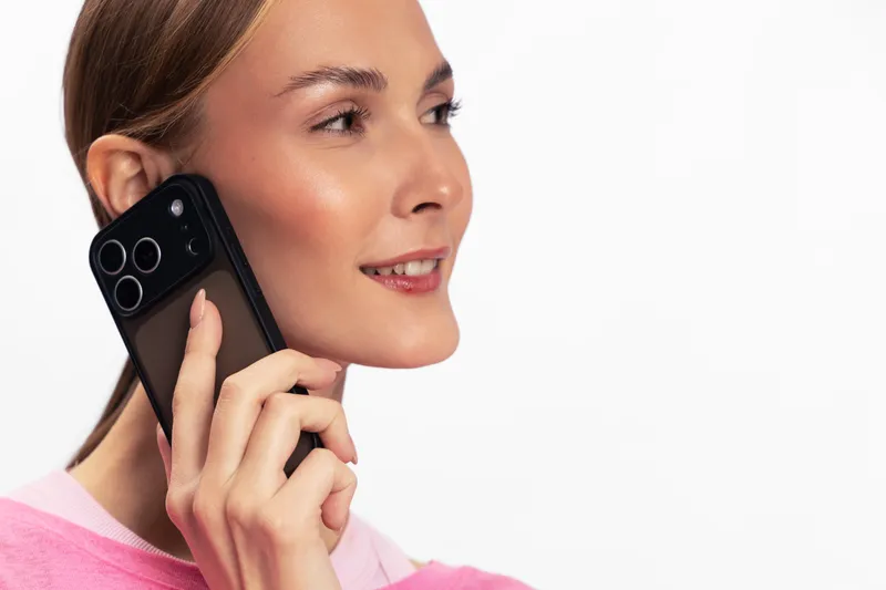 Young woman in pink sweater holds smartphone near her ear, smiling with side glance. Isolated on white background. Concept of communication, technology, and casual conversation.