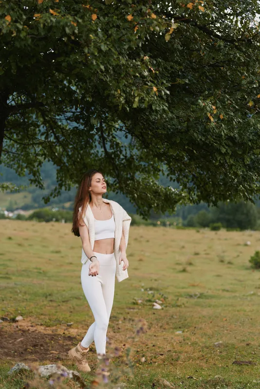 A young woman in white casualwear stands peacefully under a large tree in a green field, enjoying nature and fresh air. Concept for lifestyle, relaxation, and outdoor wellness.