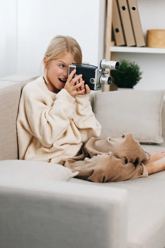 Authentic candid scene of a girl at home, playing photographer with a vintage camera, cozy sweater, soft light, and a relaxed lifestyle moment.