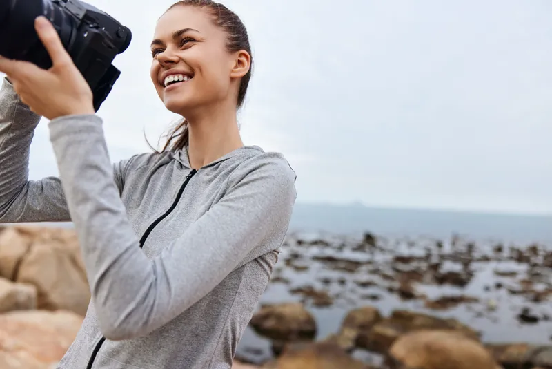 Young woman enjoying photography by the ocean, wearing a gray hoodie, showcasing happiness with a scenic background of rocks and water