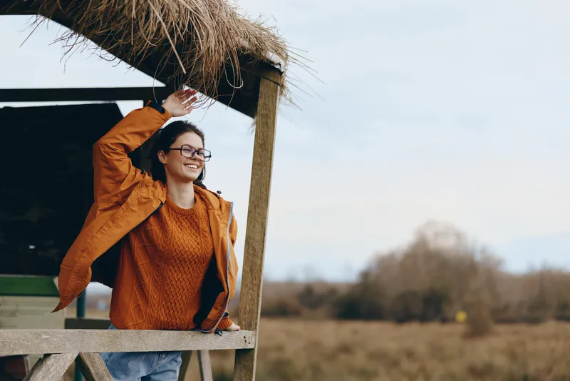 Woman lifestyle outdoors, smile under straw roof, orange jacket and casual jeans, warm sunlight, relaxed pose on wooden fence, rural field background, calm mood, daytime fresh air