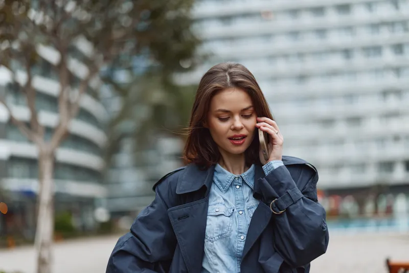 Smiling woman in a denim jacket standing outdoors in an urban street, talking on a phone. Casual fashion and city life in bright natural light, candid mood.