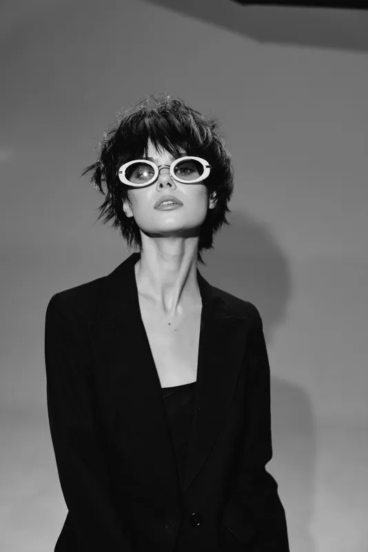 Stylish woman with short hair wearing white sunglasses in black and white portrait with dramatic lighting.