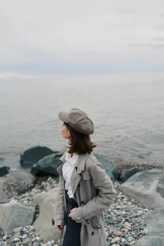 Woman in grey coat and hat stands on rocky shore looking at calm sea under cloudy sky. Concept of autumn fashion, solitude, and peaceful coastal landscape.