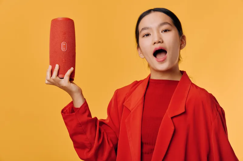 Woman holding red portable speaker with open mouth on yellow background. Young female in red jacket demonstrating wireless audio device for music and sound enjoyment.