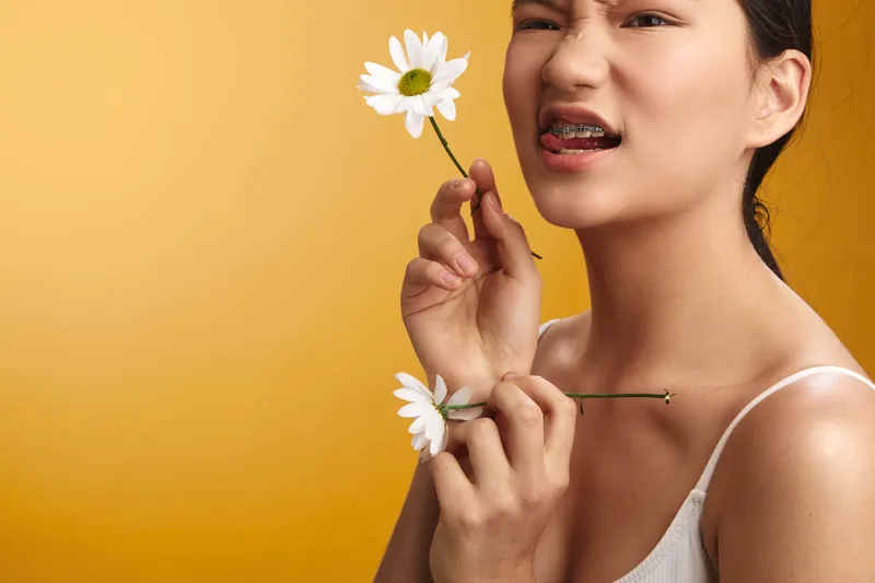 asian woman holds a delicate flower near her face, smiling softly against a warm yellow backdrop, conveying a fresh and gentle mood that highlights natural beauty and calm confidence.