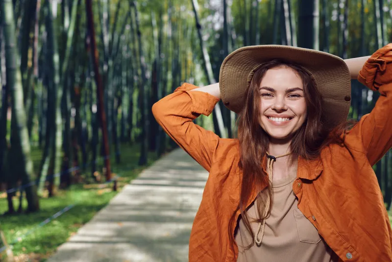 Woman smile hat park path forest portrait nature happy outdoor scene with a young woman in an orange jacket, casual lifestyle portrait on a tree-lined walkway enjoying fresh air and sunlight.