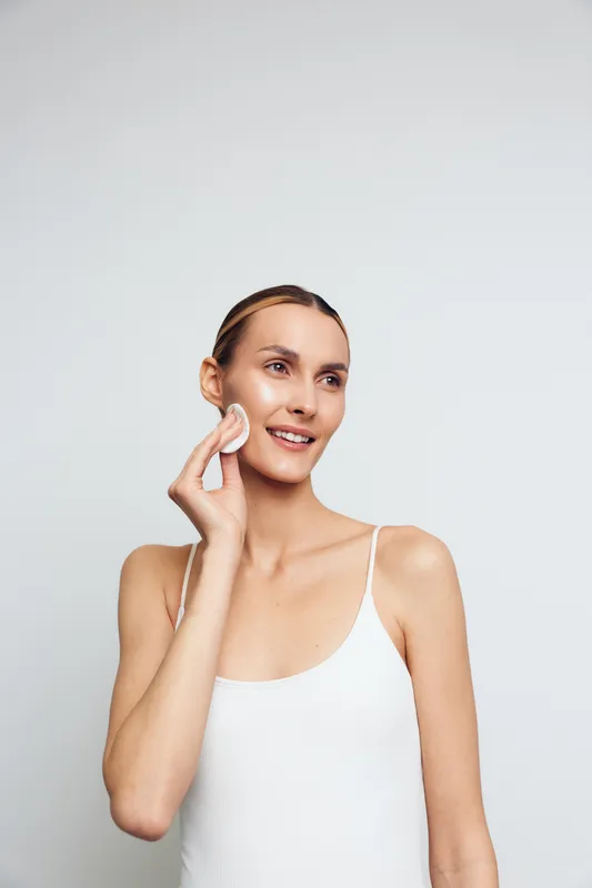 A young woman in a white tank top gently cleanses her face with a cotton pad against a plain light background. Concept for skincare, beauty routine, and natural care.