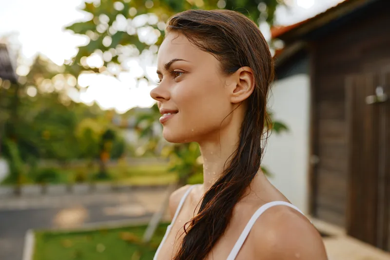 Woman portrait outdoor with wet hair, wearing a white strap top, calm side profile, summer sunlight, casual lifestyle scene, natural expression, tranquil garden background, candid and serene mood