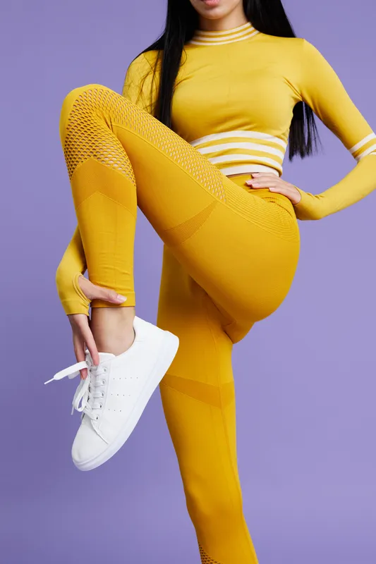 Active woman sport fitness wearing vibrant yellow seamless workout clothing stretches leg in studio with purple background, showing flexibility and strength in fitness routine.